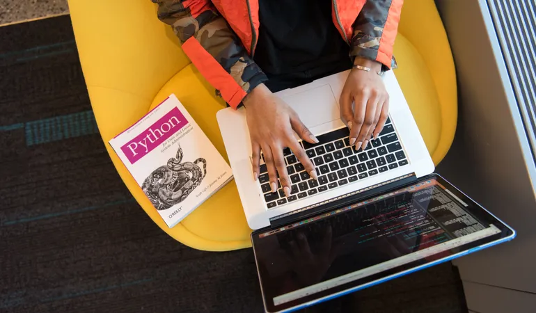 Top down view of someone using a laptop with a Python book next to them.